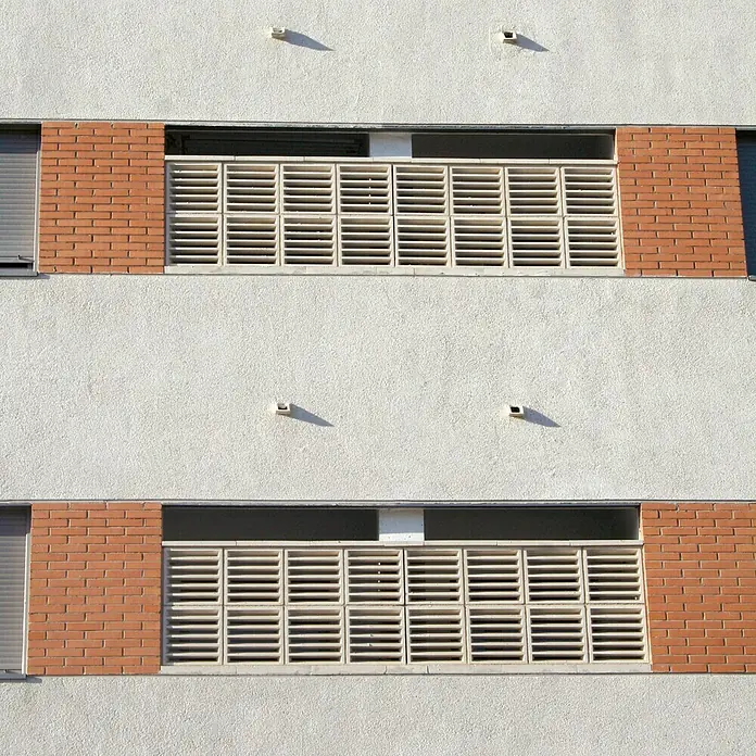 Estor plisado celular de ventilación - 40 x 40 cm, Blanco - Detalle de fachada con aberturas rectangulares con lamas horizontales, enmarcadas por ladrillos rojos bajo la luz del sol brillante.