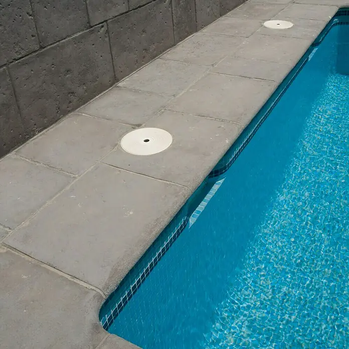Pavimento remate Piscina Opus Roca - 60 x 50 cm, Gris - La imagen muestra el borde de la piscina con losas cuadradas de piedra y cubiertas redondas blancas. El agua turquesa brilla invitando.