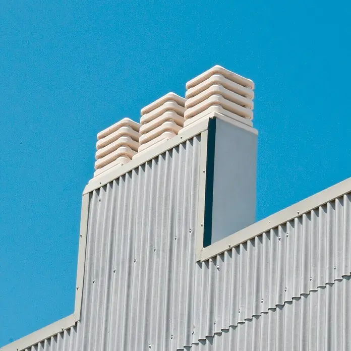 Aspirador estático 50 - Blanco - Tres chimeneas de escape en un techo de metal corrugado gris contra un cielo azul brillante. Sistema de ventilación industrial.