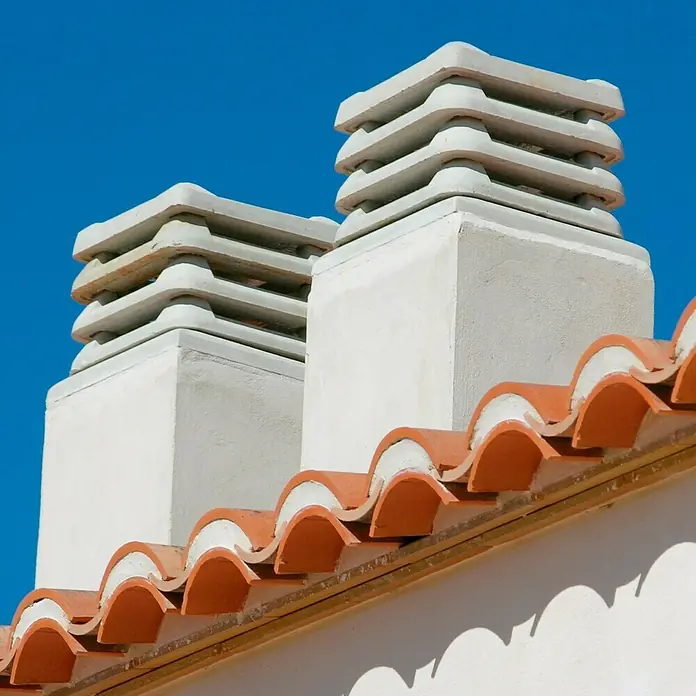 Aspirador estático 50 - Blanco - Primer plano de dos chimeneas blancas con tejas de terracota en un tejado, con un cielo azul de fondo.