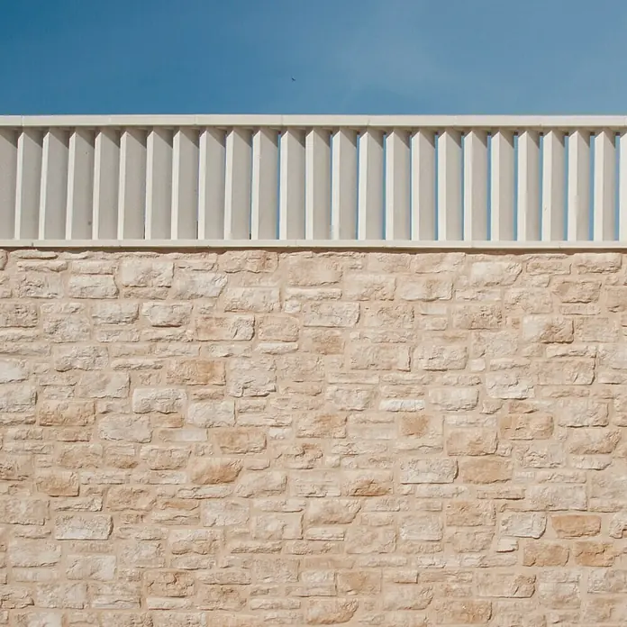 Tapa derecha de protección contra viento - Hormigón polímero, Blanco - Primer plano de una pared de piedra beige, coronada con una valla blanca y cielo azul.