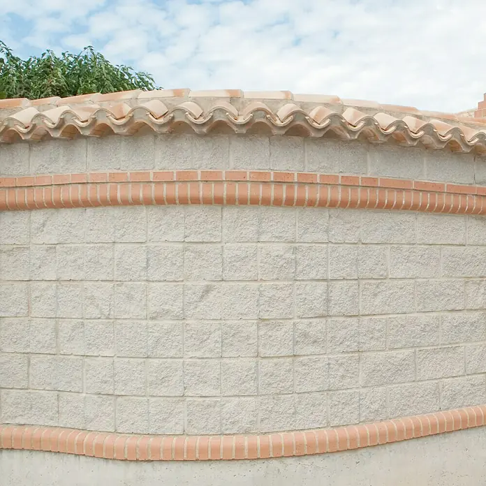 Bloque de hormigón poroso Split - 20 x 20 x 20 cm, Blanco, Hormigón, Ligeramente irregular - Imagen exterior de una pared de piedra decorativa con detalles de terracota y un techo de tejas tradicional bajo un cielo parcialmente nublado.