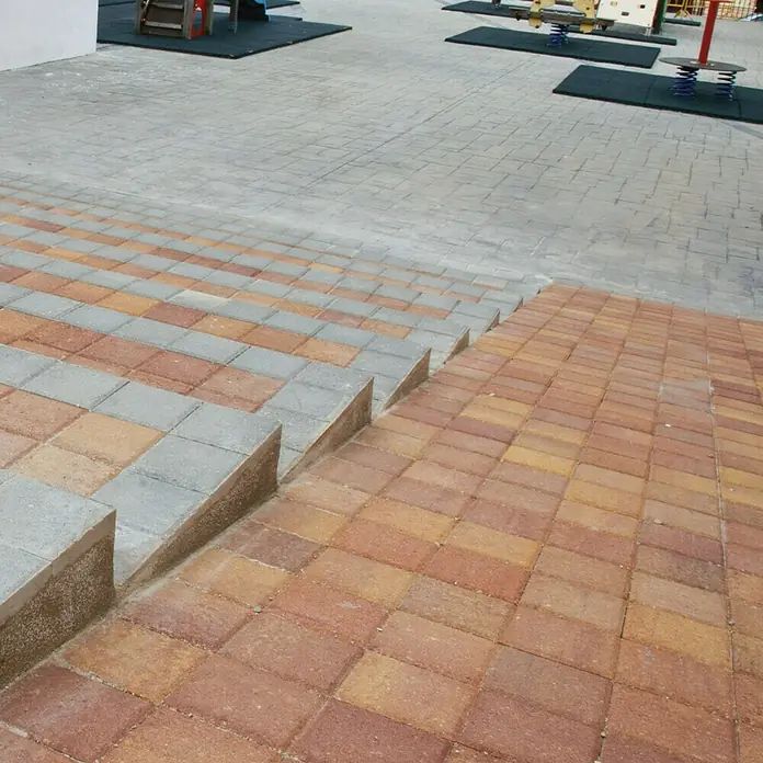 Loseta de exterior Irisada - 15 x 15 x 3,5 cm, Terracota, Hormigón - Escalones pavimentados conducen a un parque infantil. Las piedras son grises y rojas y forman un patrón atractivo.