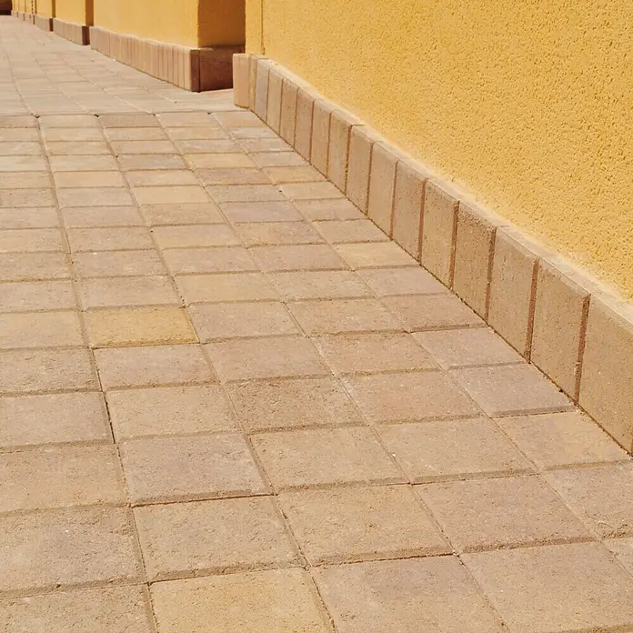 Loseta de exterior Irisada - 15 x 15 x 3,5 cm, Terracota, Hormigón - Primer plano de una pasarela pavimentada hecha de adoquines marrones junto a una pared amarilla, mostrando el diseño robusto pero estético.