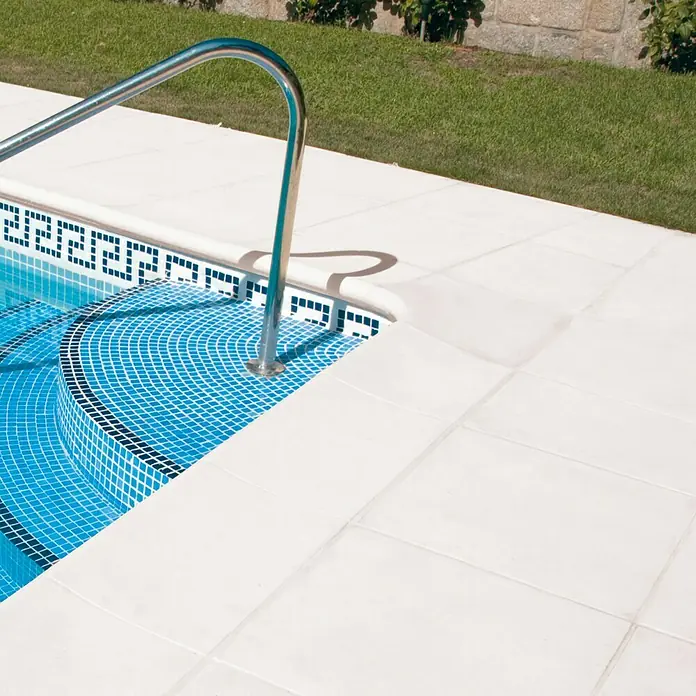 Pavimento remate Piscina - 50 x 34 cm, Blanco - Detalle de una piscina con mosaico azul y barandilla plateada, rodeada de baldosas blancas y césped verde.