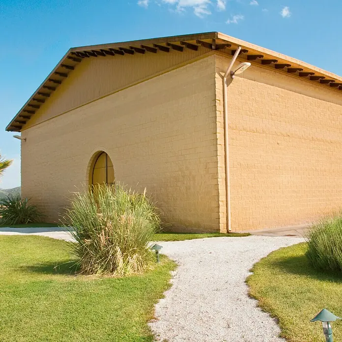 Bloque de hormigón poroso Split - 40 x 20 x 20 cm, Crema, Hormigón, Ligeramente irregular - Fotografía exterior de un edificio de ladrillo beige con una ventana redonda, rodeado de exuberante vegetación y un camino de grava bajo un cielo azul.