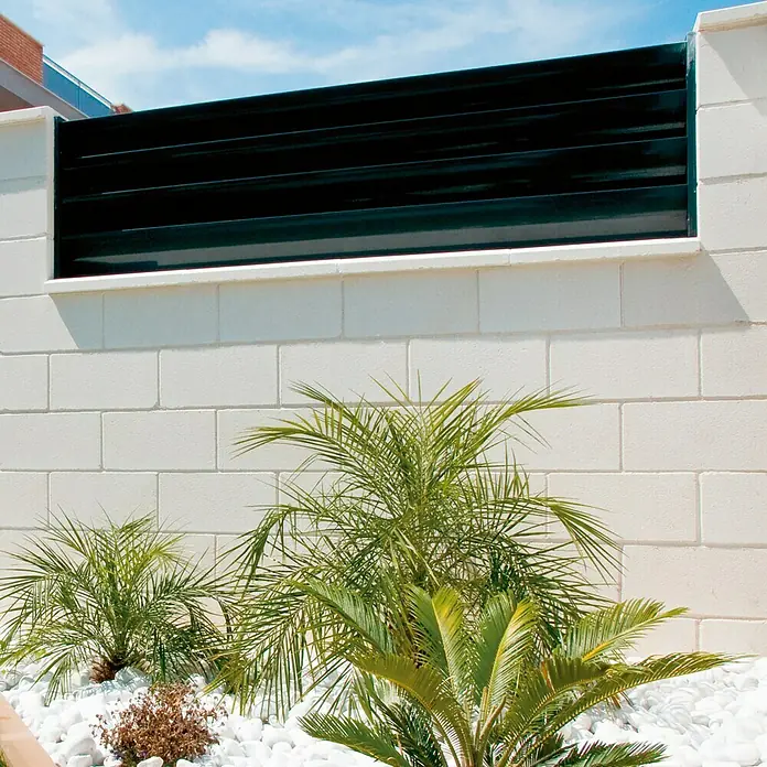 Bloque de hormigón poroso JM - 40 x 15 x 20 cm, Blanco, Hormigón - Imagen de una celosía de valla negra moderna sobre una pared blanca, rodeada de guijarros blancos y palmeras pequeñas.