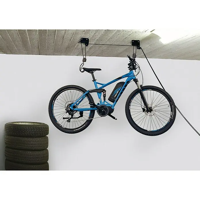Fischer Fahrradlift ProfiPlus - Traglast: 57 kg, Passend für: Gerätschaften bis 57 kg - Ein blaues Fischer E-Mountainbike hängt an einem Deckenliftsystem, während ein Stapel Reifen daneben steht.