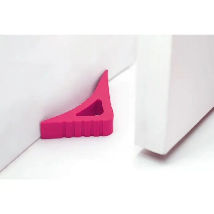 Retenedor para puerta y ventana Blockystar - Rosa - Un tope de puerta triangular rosa mantiene una puerta blanca abierta para evitar que se cierre de golpe. El producto es robusto y visible.