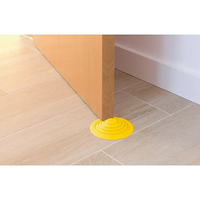 Tope de puerta y ventana Ovni Blockystar - Plástico, Ø x Al: 12 x 3 cm, Amarillo - Un tope de puerta redondo de goma amarilla mantiene una puerta abierta sobre un suelo de baldosas claras.