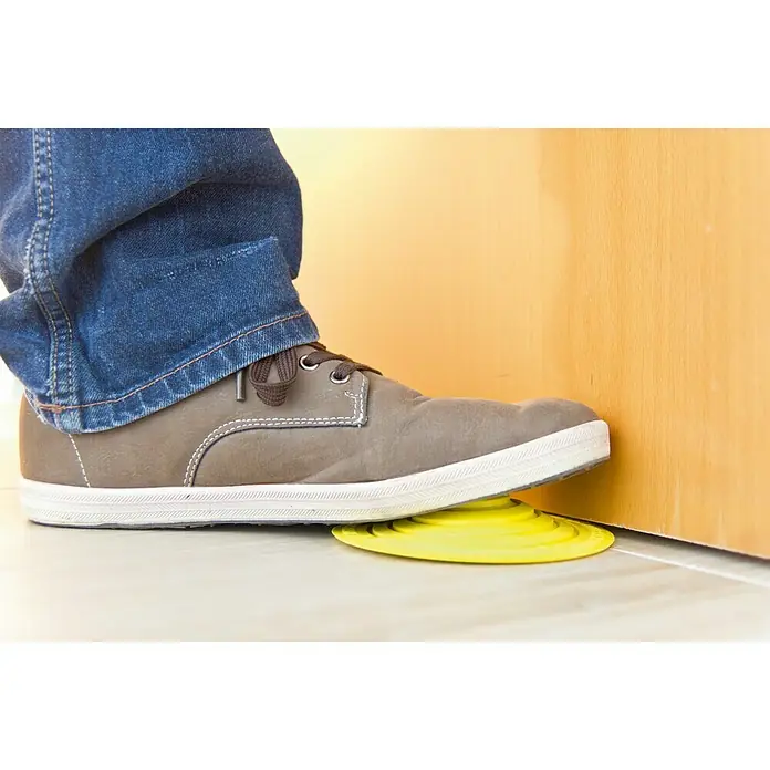 Tope de puerta y ventana Ovni Blockystar - Plástico, Ø x Al: 12 x 3 cm, Amarillo - La imagen muestra un pie presionando un tope de puerta amarillo y redondo contra una puerta para mantenerla abierta.