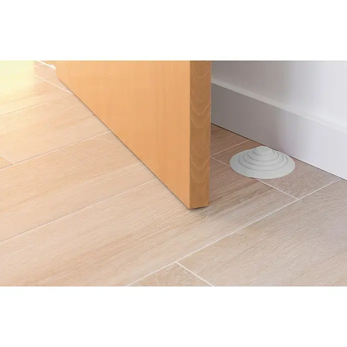 Tope de puerta y ventana Ovni Blockystar - Plástico, Ø x Al: 12 x 3 cm, Blanco - Un tope de puerta de goma gris de varios niveles mantiene una puerta de madera abierta sobre un suelo de baldosas claras.