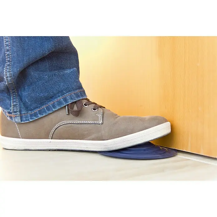 Tope de puerta y ventana Ovni Blockystar - Plástico, Ø x Al: 12 x 3 cm, Azul oscuro - Un pie con un zapato gris sujeta un tope de puerta azul para mantener una puerta abierta. Accesorio práctico para el día a día.