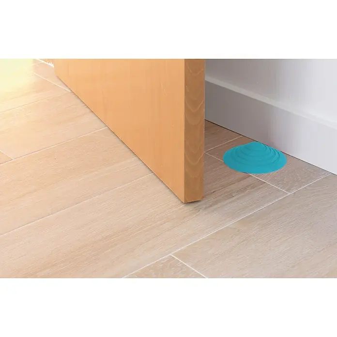 Tope de puerta y ventana Ovni Blockystar - Plástico, Ø x Al: 12 x 3 cm, Azul - Un tope de puerta de goma turquesa mantiene una puerta de madera clara abierta sobre un suelo de baldosas.