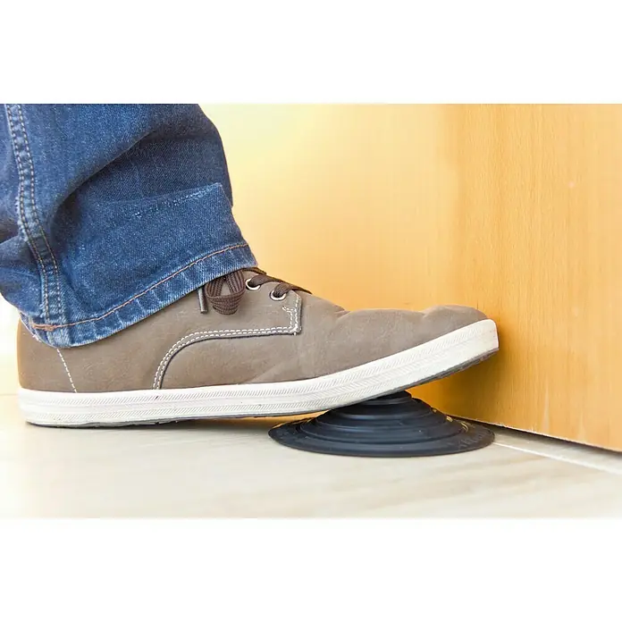 Tope de puerta y ventana Ovni Blockystar - Plástico, Ø x Al: 12 x 3 cm, Negro - Un pie sosteniendo una puerta abierta con un tope de puerta negro y flexible. El tope de puerta evita que la puerta se cierre de golpe.