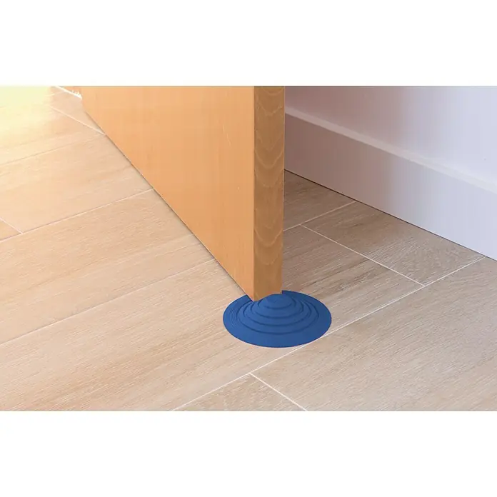 Tope de puerta y ventana Ovni Blockystar - Plástico, Ø x Al: 12 x 3 cm, Azul oscuro - Primer plano de un tope de puerta azul debajo de una puerta de madera sobre un suelo de baldosas, manteniendo la puerta abierta.