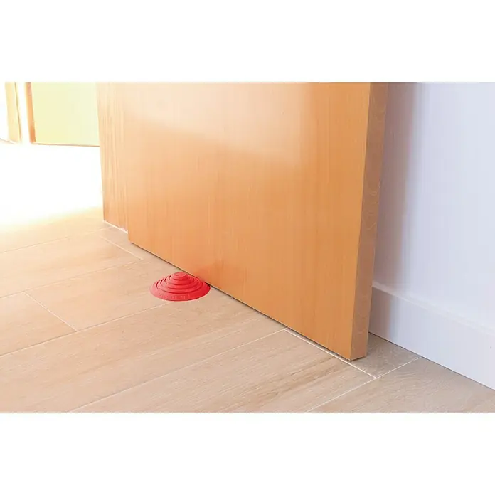 Tope de puerta y ventana Ovni Blockystar - Plástico, Ø x Al: 12 x 3 cm, Rojo - Tope de puerta cónico de silicona roja que mantiene una puerta de madera abierta sobre un suelo de baldosas claras.