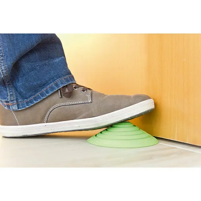 Tope de puerta y ventana Ovni Blockystar - Plástico, Ø x Al: 12 x 3 cm, Verde - Un zapato gris está pisando un tope de puerta verde en forma de cono que se coloca contra una puerta de madera para mantenerla abierta.