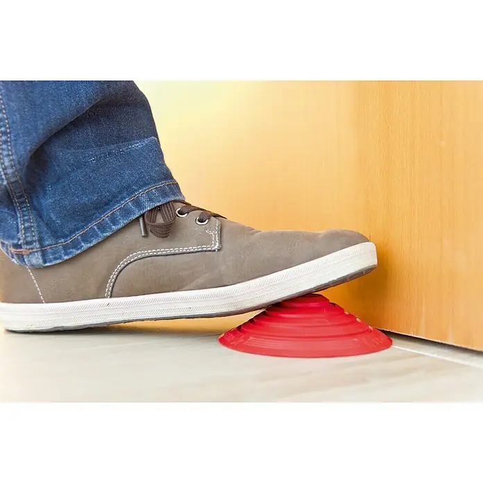 Tope de puerta y ventana Ovni Blockystar - Plástico, Ø x Al: 12 x 3 cm, Rojo - Un zapato gris presiona un tope de puerta rojo en forma de cono contra una puerta sobre un piso claro, manteniéndola abierta.