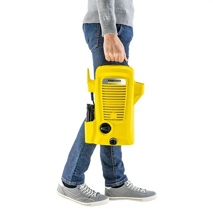 Kärcher Hidrolimpiadora de alta presión K 2 Universal Edition - 110 bar, 1.400 W, Capacidad de bombeo: 360 l/h - Una persona lleva una hidrolimpiadora amarilla Kärcher. La limpiadora es compacta e ideal para tareas de limpieza al aire libre.