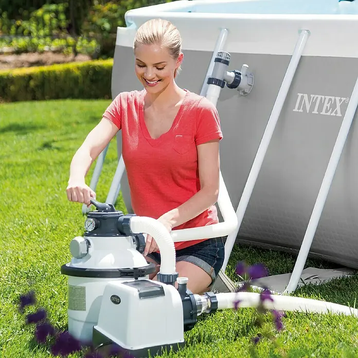 Intex Filtro de arena con panel digital - Específico para piscinas: 18.000 l, Potencia de filtración: 4.000 l/h - Una mujer opera una bomba de filtro de arena Intex junto a una piscina Intex al aire libre para mantener el agua limpia.