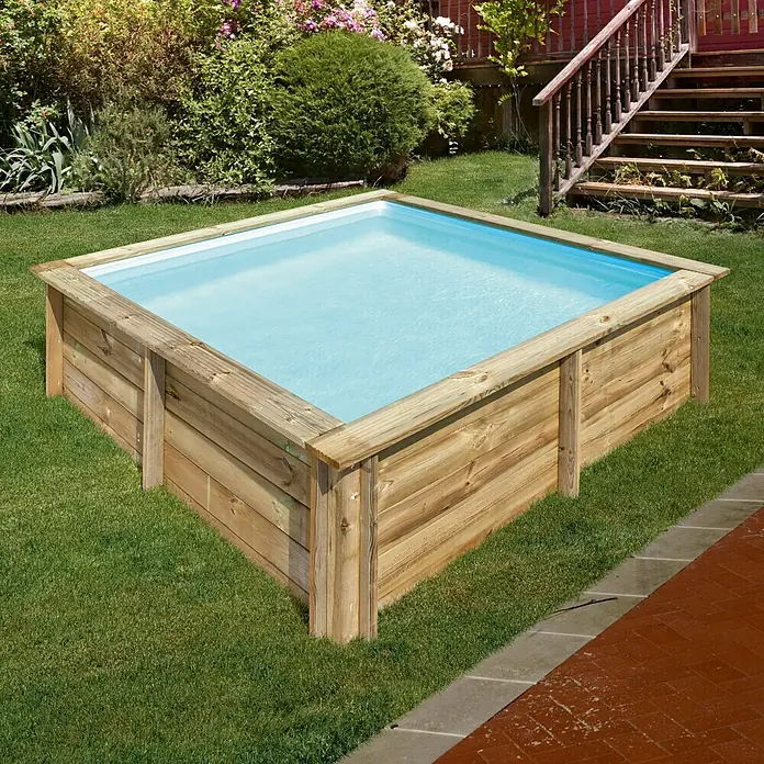Gre Holz-Pool City - L x B x H: 225 x 225 x 68 cm, Braun, 2.300 l - Gartenszene mit einem rechteckigen Holzpool im Rasen, umgeben von Grünpflanzen und einem Haus mit Treppe im Hintergrund.