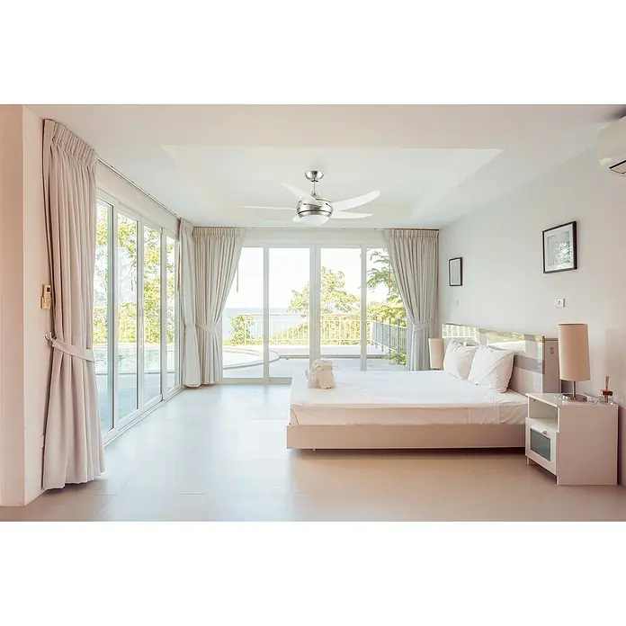 Arte confort Ventilador de techo Bela - Níquel mate, 107 cm, 18 W - Un moderno ventilador de techo con iluminación destaca en un dormitorio luminoso con ventanas panorámicas y vistas al mar.