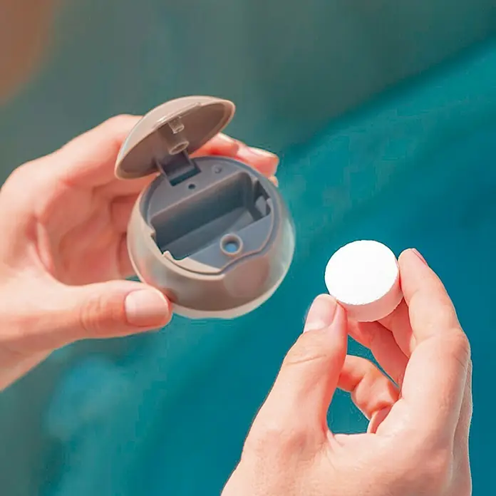 Intex Bola dosificadora de pastillas de cloro - Diámetro: 6 cm, Marrón - Las manos de una persona sosteniendo una tableta de cloro para piscinas y una cápsula dosificadora delante de un fondo azul.