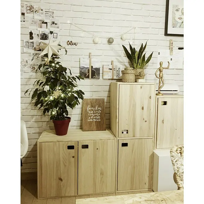 Astigarraga Dinamic Holz-Mehrzweckschrank - L x B x H: 33 x 70,8 x 53,5 cm, Kiefernholz - Wohnzimmerdekoration mit Holzschränken, Pflanze, Stern und Wanddekorationen an einer weißen Backsteinmauer.
