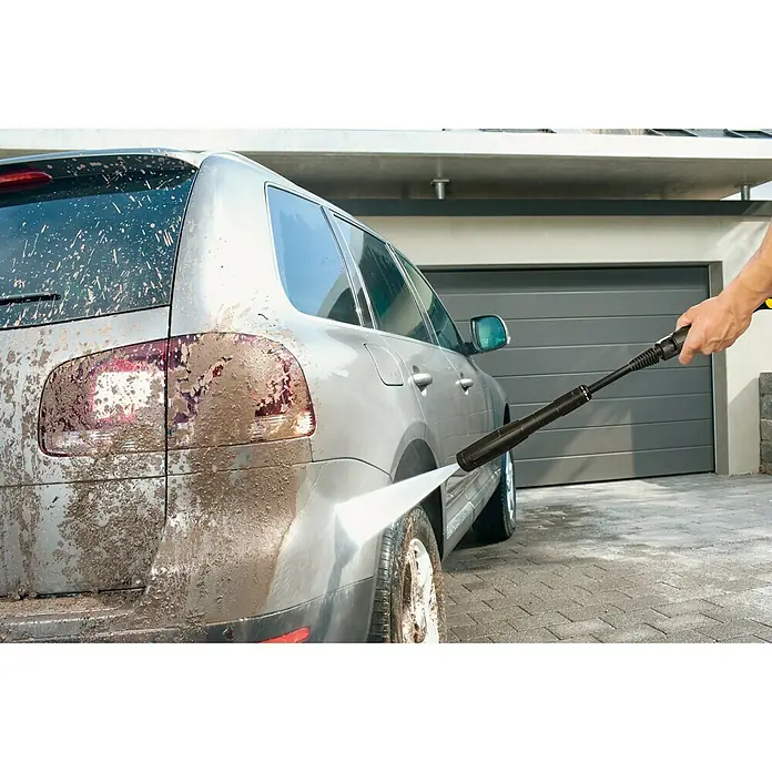 Kärcher Hochdruckreiniger K 4 Universal EditionBetriebsdruck: 110 bar - Ein Mann reinigt mit einem Hochdruckreiniger ein schlammiges Auto vor einer Garage, wobei der Fokus auf der Reinigungskraft des Geräts liegt.