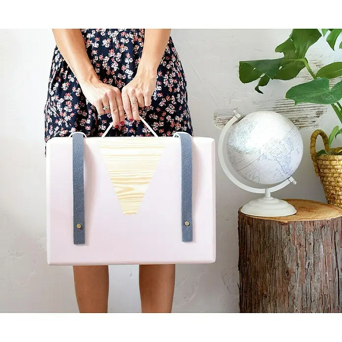 Astigarraga Caja de almacenaje - L x An x Al: 60 x 40 x 23 cm - Primer plano de una persona que sostiene un maletín rosa con detalles de madera y correas grises, con un globo terráqueo y una planta de interior cerca.