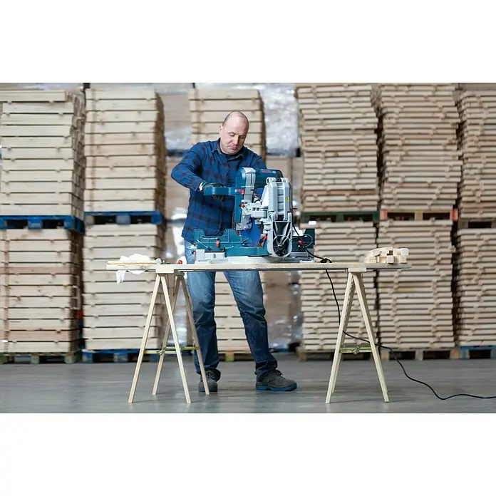 Astigarraga Caballete plegable de madera Kipli - Peso máximo admitido: 250 kg, Altura: 73,5 cm, Pino - Un hombre opera una sierra ingletadora Bosch GCM 8 SJL sobre una mesa de madera en un almacén.