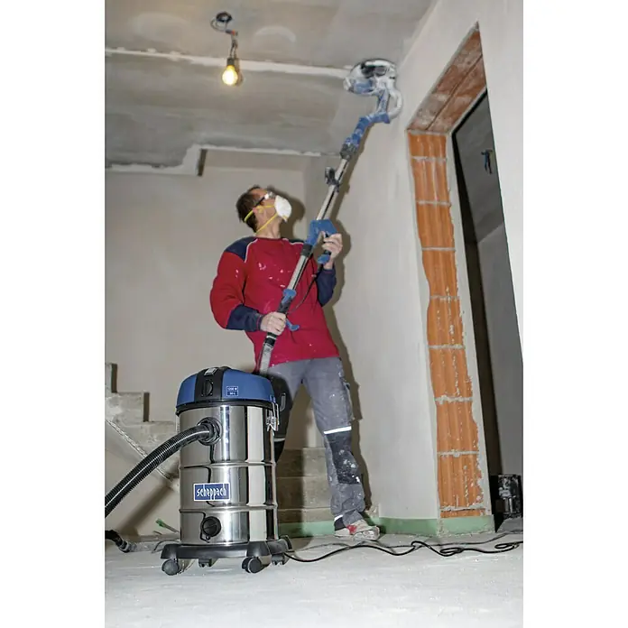 Scheppach Aspirador en seco y húmedo eléctrico NTS30 - 1.300 W, 30 l - Un trabajador utiliza una aspiradora industrial Scheppach y una lijadora para trabajar en una pared en un espacio interior.
