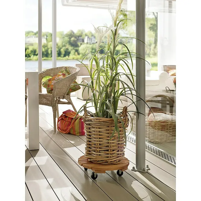 Wagner System Pflanzenroller Florenz - Durchmesser: 37,5 cm, Kiefer - Ein Korbpflanzer mit Rädern, der ein hohes Gras beherbergt, steht auf einer hellen Terrasse, ergänzt durch einen Korbstuhl und eine gestreifte Tasche.