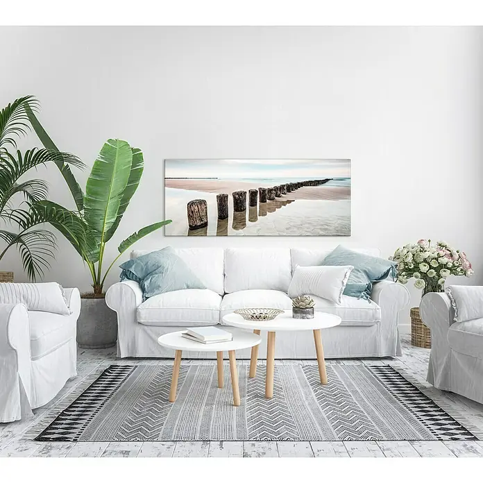 Glasbild - Strand, B x H: 125 x 50 cm - Wohnzimmer mit weißer Couch, Couchtischen und einem Teppich. Das Wandbild zeigt einen Strand und Holzpfosten im Meer.