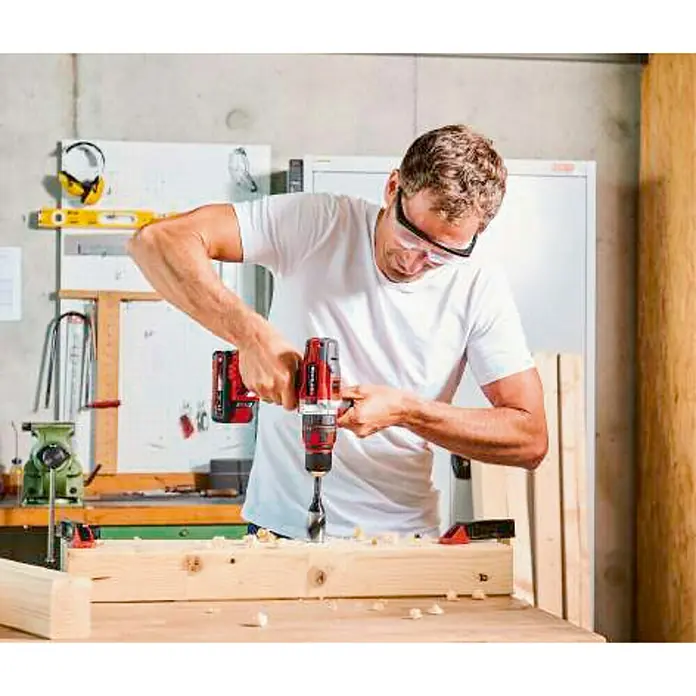 Einhell PXC 18V Taladro atornillador percutor de batería TE-CD 18/48 Li-i - 18 V, 2 baterías, 2 Ah, 48 Nm - Un hombre perfora una tabla de madera con un taladro inalámbrico Einhell mientras usa gafas protectoras.