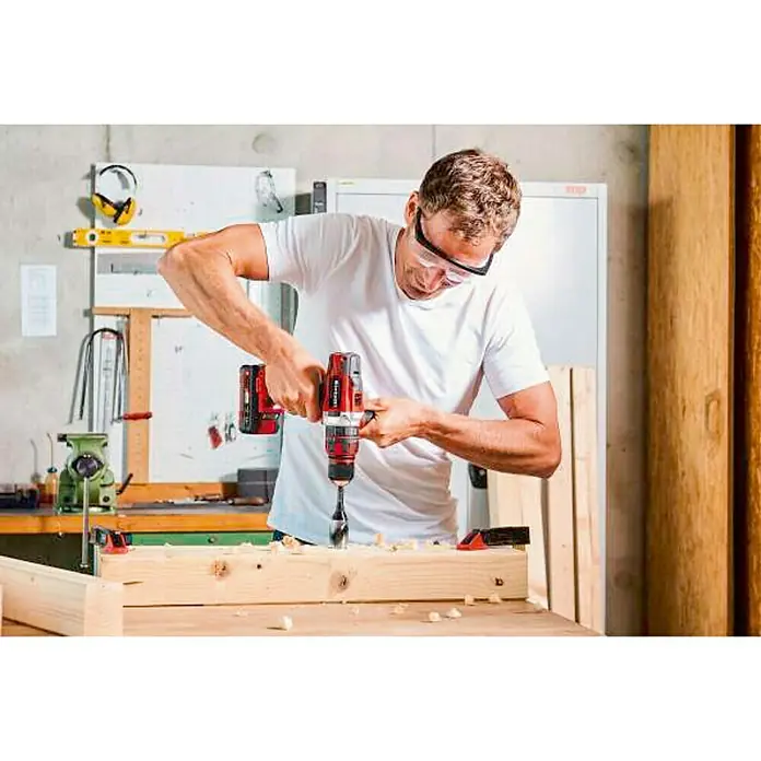 Einhell PXC 18V Taladro atornillador percutor de batería TE-CD 18/48 LI-I-Solo - 18 V, Sin batería - Un hombre hace agujeros en un trozo de madera con un taladro inalámbrico mientras usa gafas de seguridad.