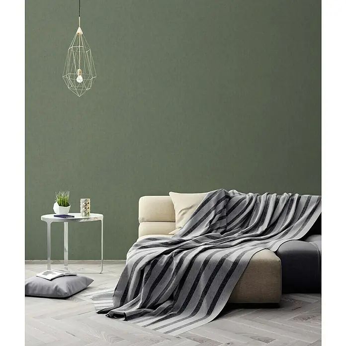 A.S. Création Geo Nordic Vliestapete - Dunkelgrün, Uni, 10,05 x 0,53 m - Wohnzimmer-Interieur mit einem beigen Sofa, das mit einer gestreiften grauen Decke bedeckt ist, und einer eleganten Drahtlampe.