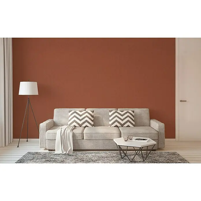 A.S. Création Geo Nordic Vliestapete - Rostbraun, Uni, 10,05 x 0,53 m - Modernes Wohnzimmer mit grauem Sofa, gemusterten Kissen, Teppich und Lampe vor einer roten Wand.