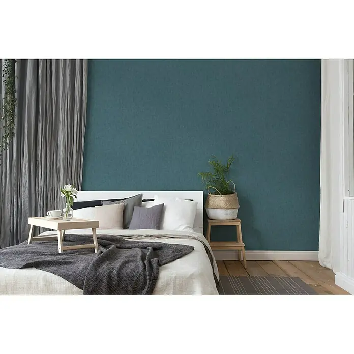 A.S. Création Geo Nordic Vliestapete - Dunkelblau, Uni, 10,05 x 0,53 m - Schlafzimmer mit einem weißen Bett, grauen Vorhängen und einer grünen Akzentwand. Das Bett verfügt über eine Ablage mit Blumen und eine Tasse.