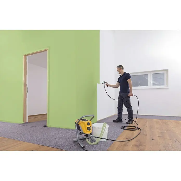 Wagner High Efficiency Airless Sistema de pulverización de pintura Airless Hea 250M - 550 W, Caudal: 1,25 l/min - El pintor utiliza un pulverizador de pintura Wagner para pintar una pared en una habitación. El dispositivo está a su lado y está conectado con una manguera.