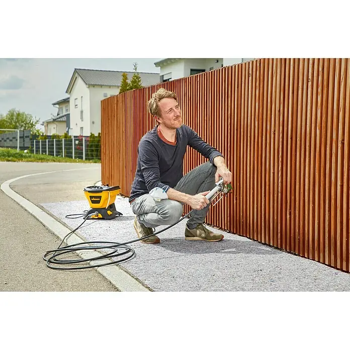 Wagner High Efficiency Airless Sistema de pulverización de pintura HEA 150M - 350 W, Caudal: 0,9 l/min - Un hombre se arrodilla y sostiene un pulverizador de pintura Wagner, pintando una cerca de madera, con el pulverizador de pintura amarillo al fondo.
