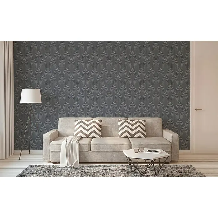 A.S. Création Pop Style Papel pintado de vellón Art Deco - Gris oscuro, Gráfico, 10,05 x 0,53 m - Escena interior con un sofá, dos cojines con estampado de galón y una lámpara de pie frente a una pared gris con diseño de plumas.