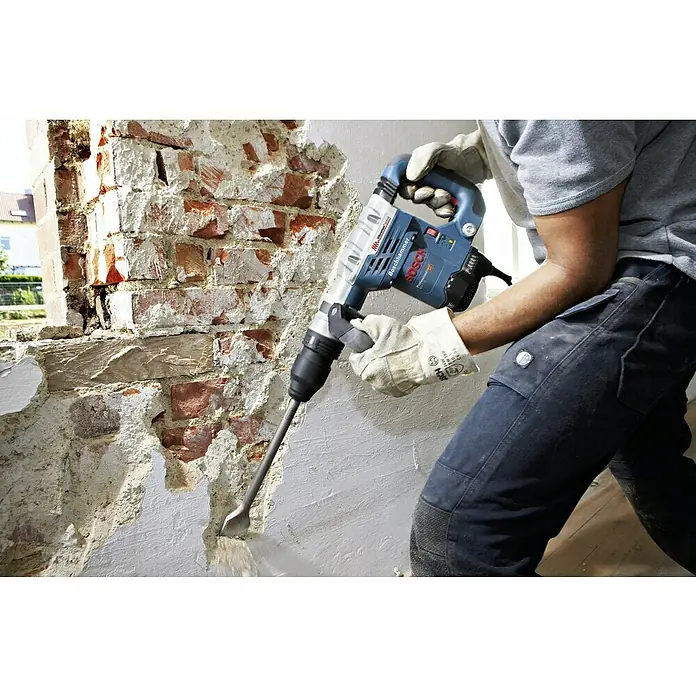 Bosch Professional Martillo demoledor GSH 5 - 1.100 W, 7,5 J, Alojamiento SDS-MAX - Imagen de un martillo rompedor Bosch en uso, rompiendo una pared de ladrillos, operado por una persona que usa guantes protectores y ropa de trabajo.