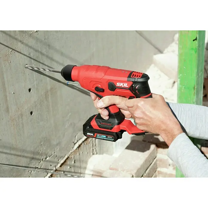 Skil PWRCore 20V Akku-Bohrhammer 3810 CA - 20 V, Ohne Akku - Der rote SKIL Bohrer wird an einer grauen Wand benutzt. Das Werkzeug ist kabellos und hat einen Akku. Das Werkzeug scheint ein Schlagbohrer zu sein.