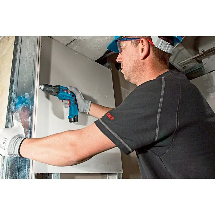Bosch Professional Atornillador para placas de yeso de batería GTB 12V-11 LBoxx - 12 V, Sin batería, Incluye maletín - Un trabajador de la construcción usa un taladro inalámbrico azul de Bosch para asegurar paneles de yeso a un marco de metal.