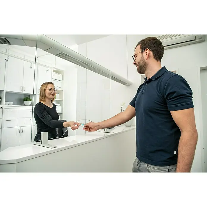 Room Plaza Hygieneschutzglas mit Durchreiche - 600 x 500 mm, Einscheibensicherheitsglas (ESG) - Ein Mann übergibt eine Karte an eine Frau hinter einem Spuckschutz aus Glas. Das Produkt ist eine Schutzvorrichtung für Theken.
