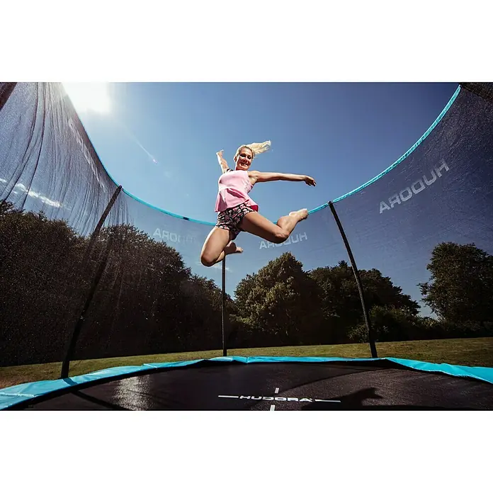 Hudora Trampolin Fantastic Complete 300 V - Durchmesser: 300 cm - Eine junge Frau springt auf einem HUDORA-Trampolin mit Sicherheitsnetz und blauem Rand vor blauem Himmel.