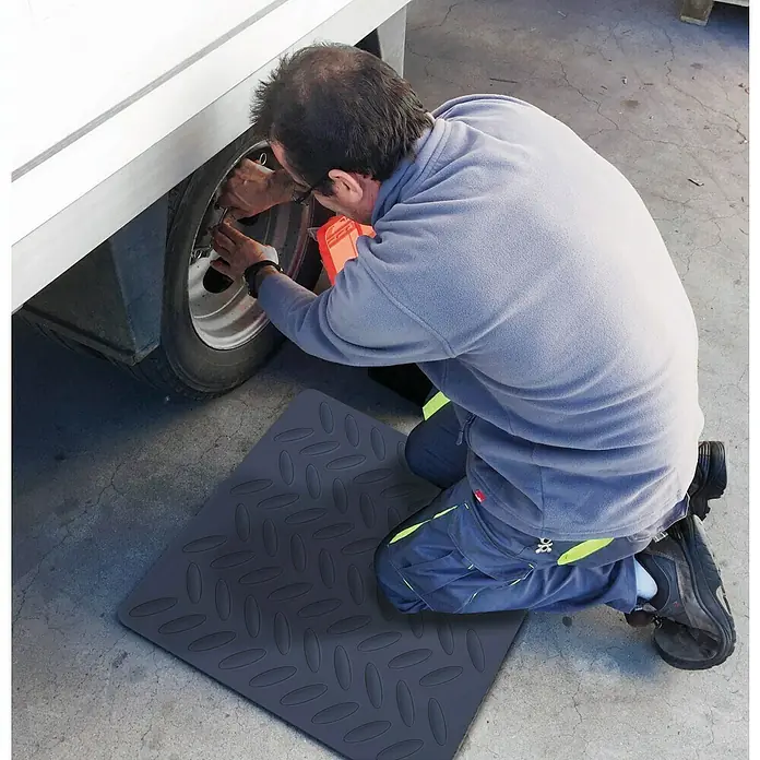 Estera antideslizante - L x An: 60 x 60 cm, Negro - Una rodillera gris está extendida en el suelo mientras un hombre trabaja en un neumático, protegiendo sus rodillas.