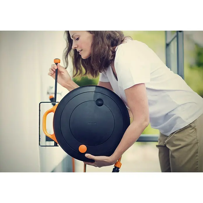 Fiskars Wand-Schlauchhalterung - Passend für: Fiskars Schlauchwagen Waterwheel S - Frau hält einen Fiskars Schlauchaufroller in den Händen. Der Schlauchaufroller ist schwarz mit orangefarbenen Akzenten und Haltegriffen.
